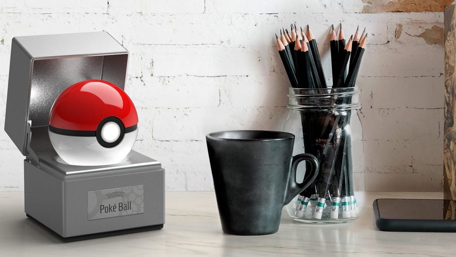 Die cast poke ball in case, beside a black cup and a clear pencil holder with several black pencils all on a white desk