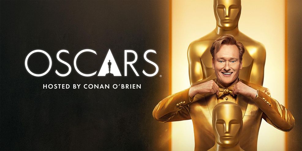 Conan O'Brien in a gold suit next to two large oscars statues